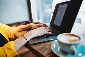 Hands of a woman wearing a yellow blouse and is typing on a laptop. To the right of the laptop, and in the foreground is a latte in a green cup.